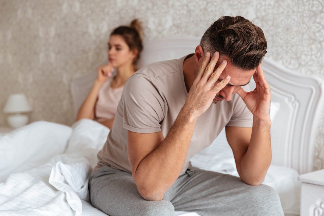 Casal na cama, homem sentado à beira, cabisbaixo